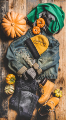 Autumn outfit layout. Flat-lay of vintage denim jacket, sweater, jeans, woolen scarf, hat, yellow boots, glasses, pumpkin, candle and tea in cup over wooden background, top view. Fall apparel and mood