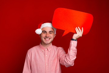 Happy young man in santa claus hat holding red speech bubble over red background
