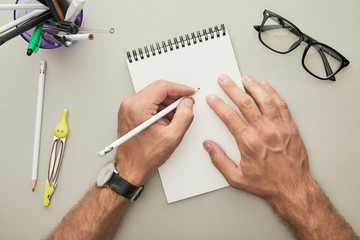 cropped view of man writing in notebook near glasses and stationery isolated on grey