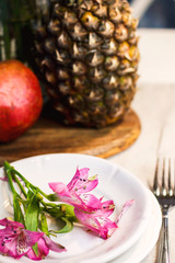 Tropical paradise heaven asian  pink iris flower on white plate, pineapple, pomegranate  in spa resort, restaurant. Holidays rest at sea. Food of gods. Table setting decoration knife
