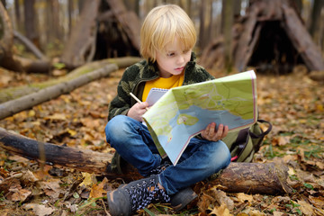 Little boy scout is orienteering in forest. Child is sitting on fallen tree and looking on map on...