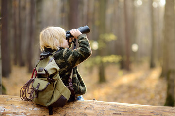 Little boy scout with binoculars during hiking in autumn forest. Child is sitting on large fallen tree and looking through a binoculars. - obrazy, fototapety, plakaty