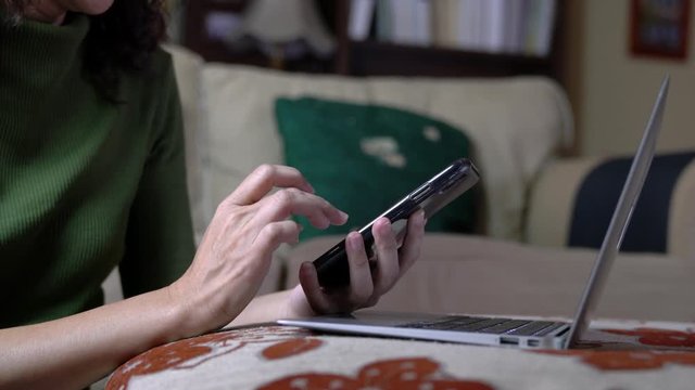 Woman surfing on her cell phone with laptop on sofa, home environment. Work from home or flexi time work concept.