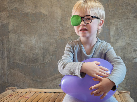 Funny blond kid in gray clothes holds purple ball in his hands. Glasses with thick glasses and green occluder