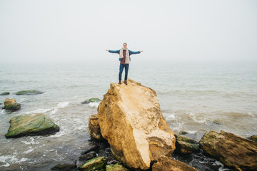Fototapeta na wymiar Traveler man stands on a rock against a beautiful sea with waves his hands to the side. Stylish hipster boy posing. Place for text or advertising