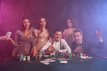Friends playing poker at casino, at table with stacks of chips, money, cards on it. Celebrating win, smiling. Black, smoke background. Close-up. - obrazy, fototapety, plakaty