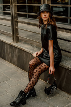 Outdoor full-length street fashion portrait of young elegant confident model, woman wearing trendy leather bucket hat, dress, black leopard print tights, lace up ankle boots, holding stylish small bag