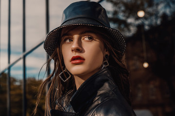 Outdoor close up fashion portrait of young beautiful confident model, woman wearing trendy faux leather bucket hat, long earrings, trench coat, posing in city street. Copy, empty space for text © Victoria Fox