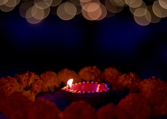 Festive black background design with tinge of blue and space for text in the middle. Illuminated red color mud lamps surrounded by orange marigold flowers with shimmering yellow blurred lights.