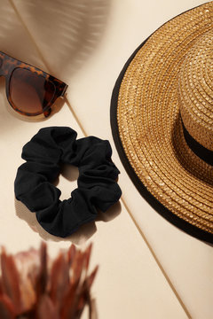 Subject shot of a black textile hair band. The scrunchie is isolated on the beige fabric surface and surrounded with a brown straw hat, sunglasses and tropic flowers. 