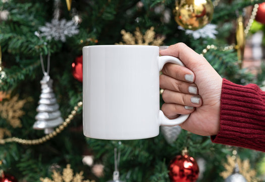women hand holding white ceramic coffee cup on christmas tree background. mockup for creative advertising text message or promotional content.