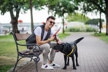 Young blind man with white cane and guide dog sitting in park in city.