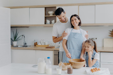 Obraz na płótnie Canvas Happy family cook together at kitchen. Father, mother and dauther busy preparing delicious meal at home. Husband embraces wife who whisks and prepares dough, bake cookies. Food, togetherness