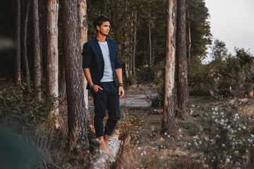 Handsome man posing outdoors in forest stands barefoot on a log, wearing checked jacket. Place for text or advertising