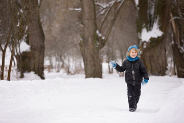 Fototapeta na wymiar Kid boy is walking along a snowy track in a park. Cheerful child with blond hair in blue mittens, a hat and a black jacket. Activities with children outdoors on a frosty winter day.