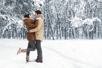 Fototapeta na wymiar Couple Embracing Standing In Snowy Winter Forest In The Morning