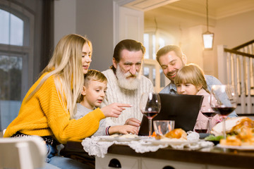 Fototapeta na wymiar Happy smiling family looking film or making call via internet using laptop, sitting at the festive tabe at home, celebrating dinner together. Thanksgiving day concept
