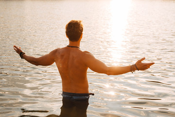 Man with perfect muscular stands in the water at sunset flinging his hands to the side. Shoot from the back. Concept of freedom relaxation. Place for text or advertising