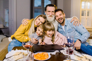 Obraz na płótnie Canvas Mom, dad and their little children, grandfather have holiday dinner , sitting at the table in cozy room, using laptop and talk. Happy family concept.