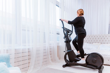 Model - a fat woman, trying to lose weight and at home is engaged in an elliptical trainer - obrazy, fototapety, plakaty