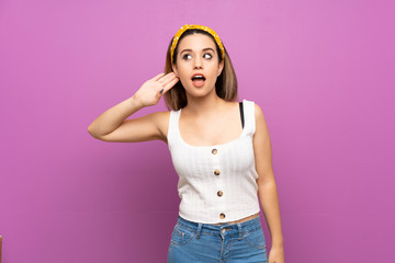 Pretty young woman over isolated purple wall listening to something by putting hand on the ear