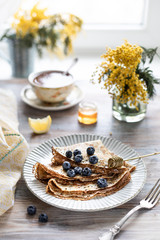 Obraz na płótnie Canvas A plate with pancakes with blueberry berries on a wooden table. In the background is a cup of tea and a bouquet of spring flowers by the window.