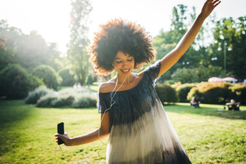 Young black woman dancing listening to the music with earphones from the smart phone in a park at...