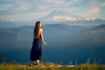 A beautiful girl in a light summer dress in the mountains of the Caucasus, admires the sunset and enjoys freedom and clean mountain air.