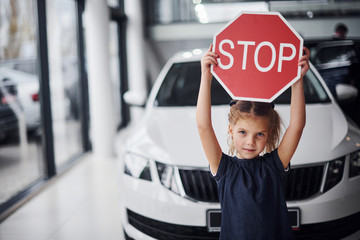 Portrait of cute little girl that holds road sign in hands in automobile salon