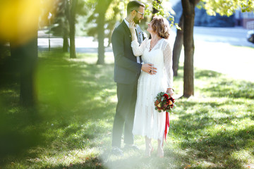 Fototapeta premium Młoda para spędza romantyczne chwile spacerując po parku. Stylowa panna młoda i pan młody pozowanie i całują się w parku w dniu ślubu. Elegancka panna młoda w pięknej białej sukni, pan młody w garniturze.