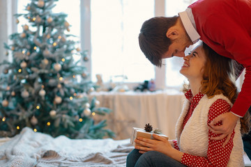 Happy loving couple enjoying Christmas. Young man touching his girlfriend nose with his nose holding a present with window on background