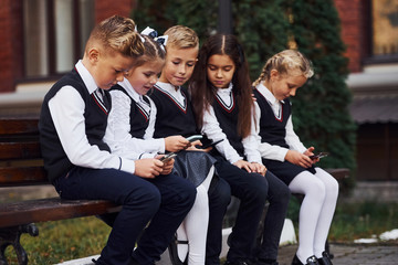 School kids in uniform that sits outdoors on the bench with smartphones