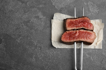 Carving fork with slices of delicious beef steak on grey table, top view. Space for text