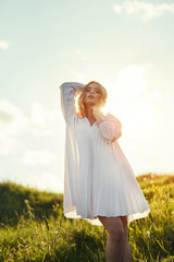 Fototapeta na wymiar Girl in a long white dress dancing in the field. Blonde woman in the sun in a light dress. Girl resting and dreaming, perfect summer makeup on her face