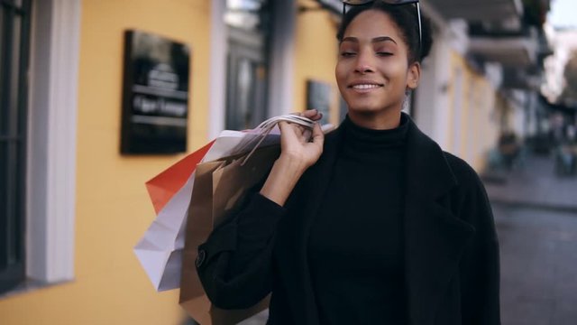 Portrait of an attractive mixed race girl smiling while walking down the street with colorful shopping bags. Happy young woman walking after shopping by fancy streets city, window shopping