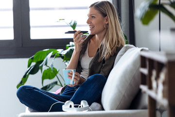 Pretty young woman talking on mobile phone with hands free while drinking cup of coffee on sofa at...