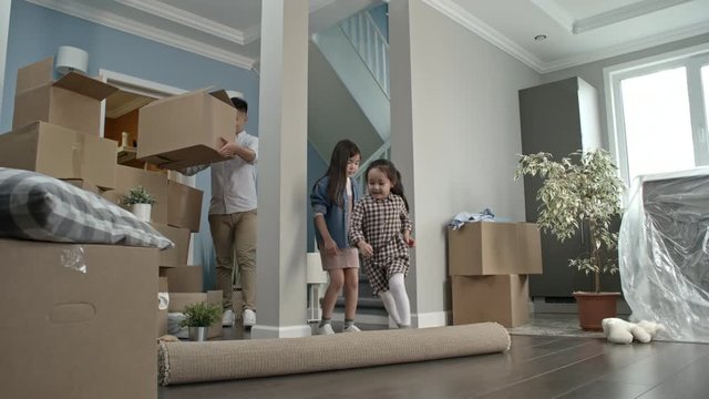 PAN of cute little girls unrolling carpet in living room of their new house as their cheerful parents unloading cardboard boxes