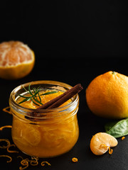 Citrus (tangerine) jam in a glass jar on a table with a cinnamon stick and a branch of rosemary. Around segments of mandarin, shavings of zest. Close-up, selective focus. Black stone background.