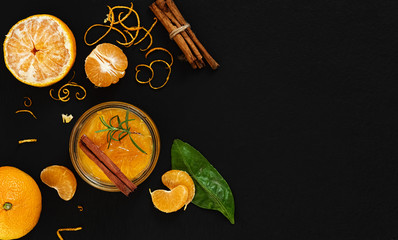 Citrus (tangerine) jam in a glass jar on a table with a cinnamon stick and a branch of rosemary. Around segments of mandarin, shavings of zest. Top view, copy space. Black stone background.