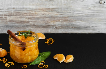 Citrus (tangerine) jam in a glass jar on a table with a cinnamon stick and a branch of rosemary. Around segments of mandarin, shavings of zest. Close-up, copy space. Black stone background.