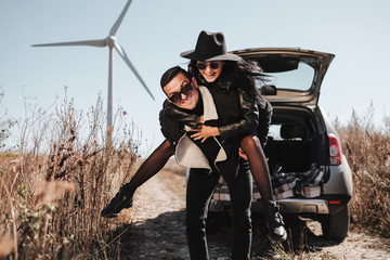 Obraz premium Happy Traveling Couple Enjoying a Car Trip on the Field Road with Electric Wind Turbine Power Generator on the Background