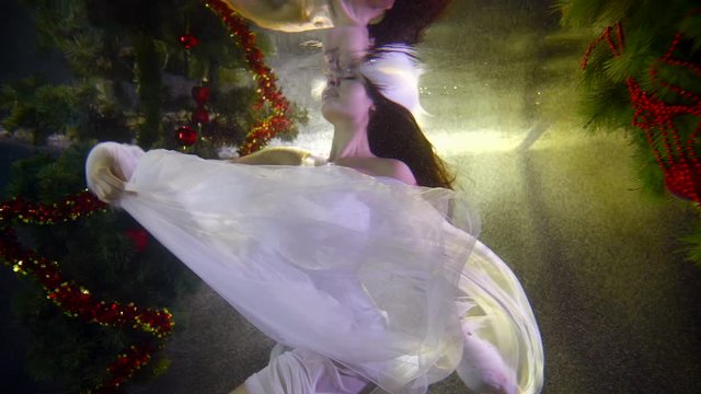 A girl in a white Christmas sexy dress that has tulle fluttering, she is underwater on a dark background with lights. around it two festive elegant Christmas trees. She makes beautiful movements.