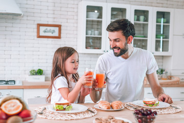 smiling father and daughter clicking glasses with drinking juice for breakfast in the kitchen - obrazy, fototapety, plakaty