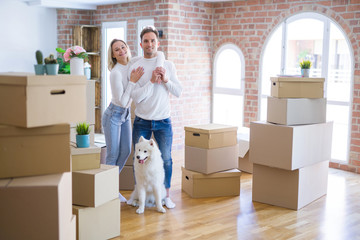 Young beautiful couple with dog standing at new home around cardboard boxes