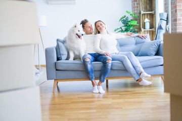 Young beautiful couple with dog sitting on the sofa at new home around cardboard boxes