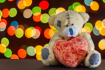Soft toy bear with heart on the background of bokeh of colorful lights of garlands. Copy space for text, selective focus