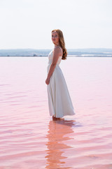 Fototapeta na wymiar Side view of cute teenager woman wearing white dress standing on an amazing pink lake
