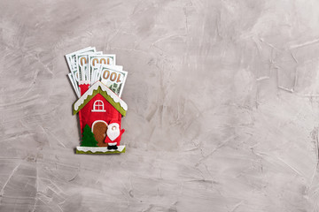 Bundle of US dollars in the funny santa claus house from felt on the gray concrete background. Cristmas Concept.