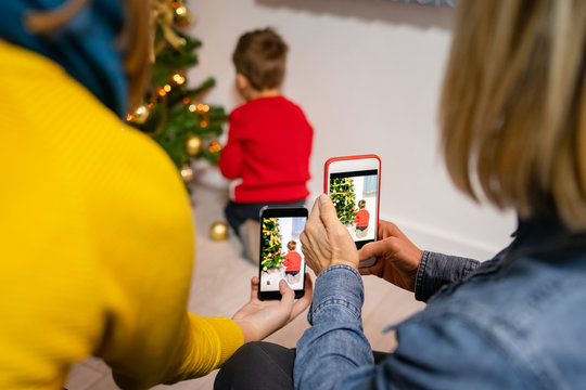Small boy decorating Christmas tree while two women taking photos or video of him with smart mobile phones mother and grandmother or aunt family parents female back view