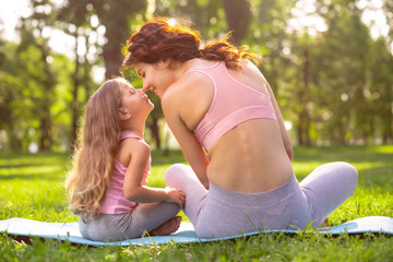mother and small daughter in sport wear sitting on the yoga mat nose to nose, back view